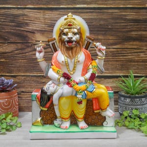 May include: A colorful statue of a Hindu deity with a lion head, sitting on a throne with a figure lying across their lap. The deity is adorned with a garland of flowers and a yellow and orange robe. The statue is made of marble and has intricate details.