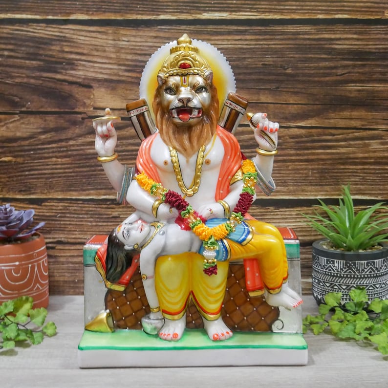 May include: A colorful statue of a Hindu deity with a lion head, sitting on a throne with a figure lying across their lap. The deity is adorned with a garland of flowers and a yellow and orange robe. The statue is made of marble and has intricate details.