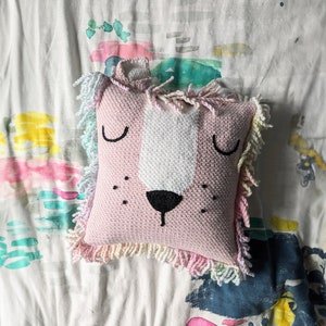 A crocheted pillow of a pink lion face on a colourful bedspread