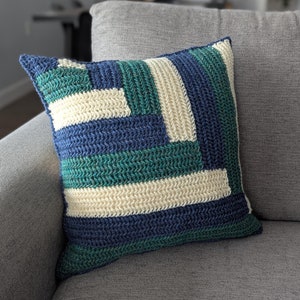 May include: A blue, green, and white crocheted pillow with a geometric pattern. The pillow is sitting on a gray couch.