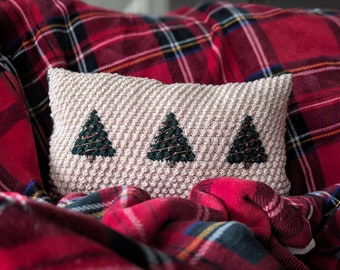 Puede incluir: Una almohada de color crema con tres árboles de Navidad bordados en verde sobre un sofá de cuadros rojos.