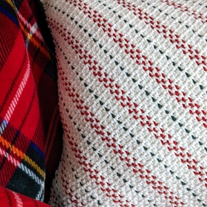 Close up of the side of a crocheted white, red and green striped pillow on a red plaid blanket, with a lit up Christmas tree in the background.