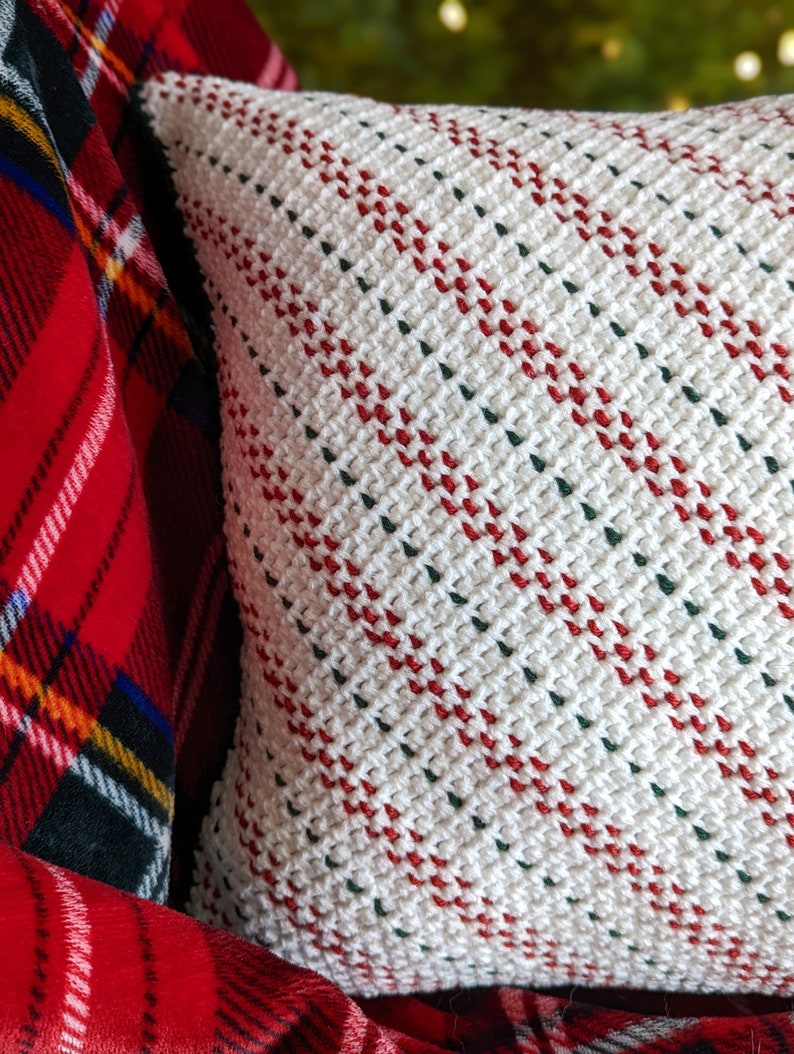 Close up of the side of a crocheted white, red and green striped pillow on a red plaid blanket, with a lit up Christmas tree in the background.