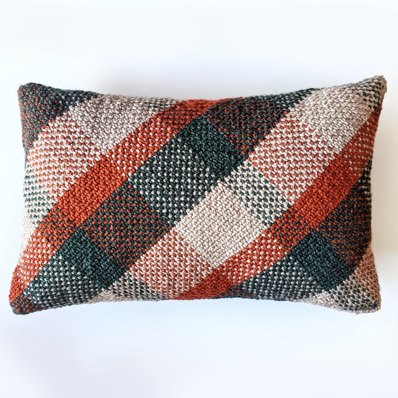 A rectangular, woven-looking pillow in burnt orange, forest green and cream colours, leaning on a white background