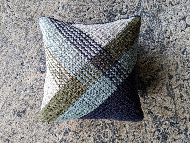 A blue, green and white crocheted pillow on a textured carpet