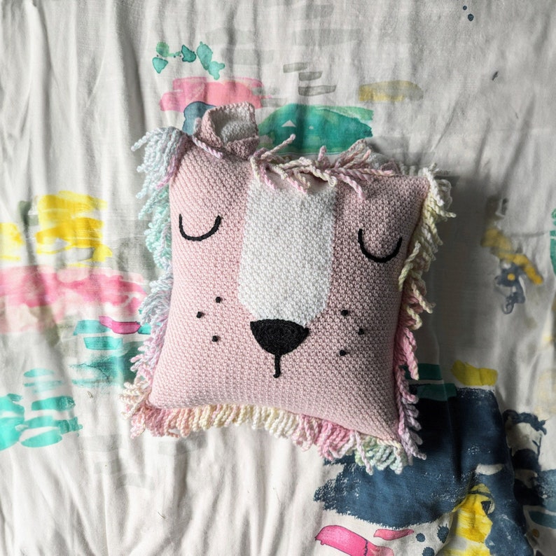 A crocheted pillow of a pink lion face on a colourful bedspread