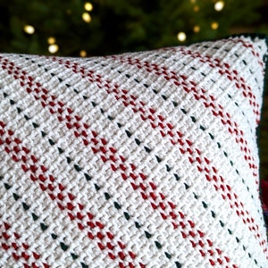 Close up of the top of a crocheted white, red and green striped pillow, with a lit up Christmas tree in the background.