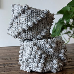May include: A handmade, gray crochet pillow with a textured, bobble stitch design. The pillow is made of a speckled yarn and sits on a wooden surface. The bobbles are evenly spaced across the surface.