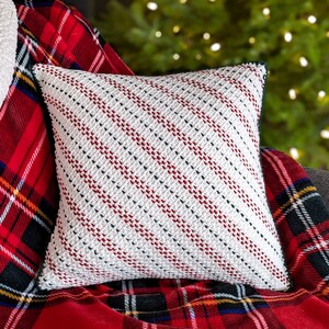 A crocheted white, red and green striped pillow on a red plaid blanket and grey couch, with a lit up Christmas tree in the background.