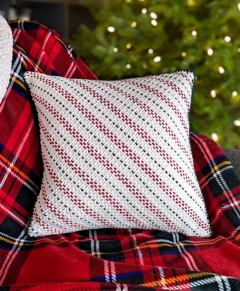 A crocheted white, red and green striped pillow on a red plaid blanket and grey couch, with a lit up Christmas tree in the background.