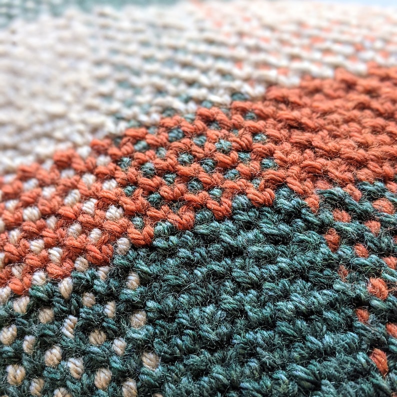 A closeup detail of a rectangular, woven-looking pillow in burnt orange, forest green and cream colours