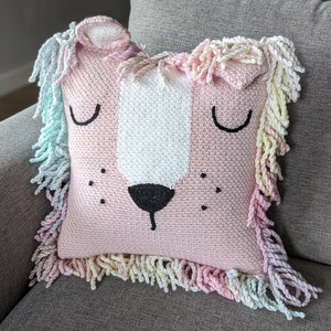 A crocheted pillow of a pink lion face on grey couch