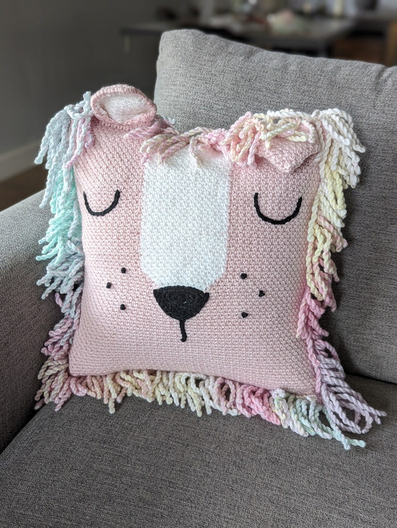 A crocheted pillow of a pink lion face on grey couch