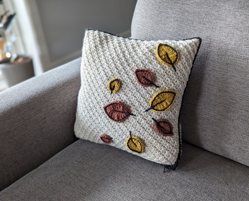 A crocheted pillow in cream-coloured yarn with red and gold leaves on a grey couch