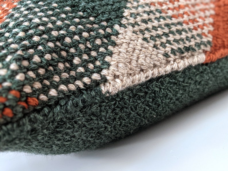 A close-up angle of a woven-looking pillow in burnt orange, forest green and cream colours.