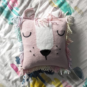 A crocheted pillow of a pink lion face on a colourful bedspread