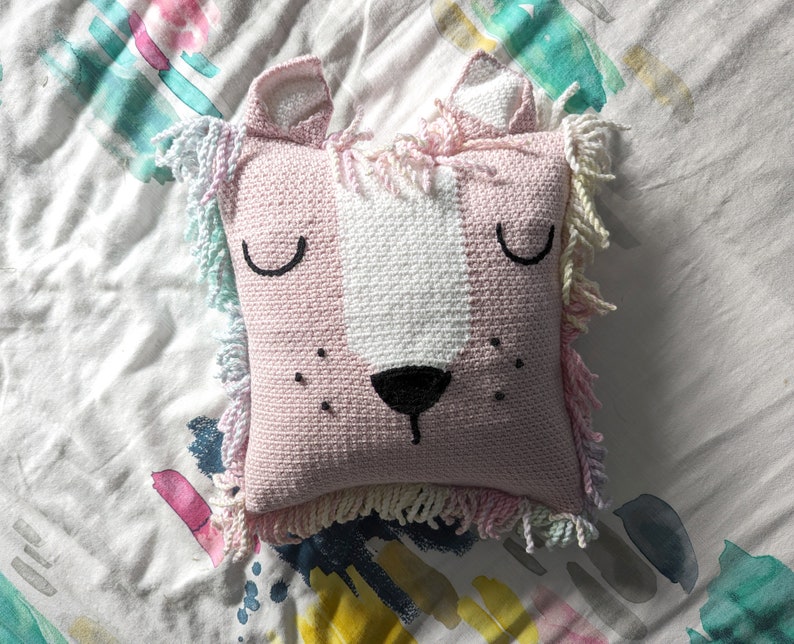 A crocheted pillow of a pink lion face on a colourful bedspread