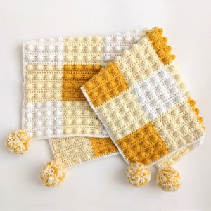 May include: A crocheted baby blanket with a checkered pattern in white and yellow. The blanket has a textured, puffy surface and is trimmed with pom-poms.