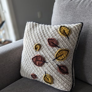 A crocheted pillow in cream-coloured yarn with red and gold leaves on a grey couch