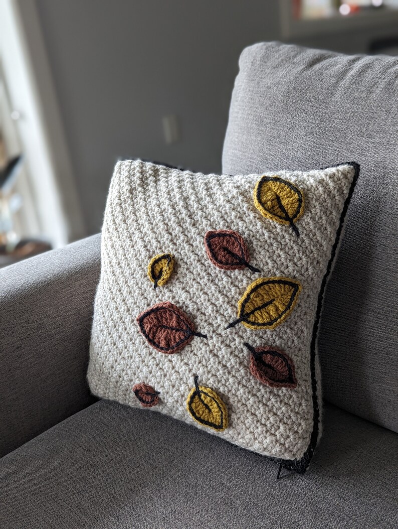 A crocheted pillow in cream-coloured yarn with red and gold leaves on a grey couch