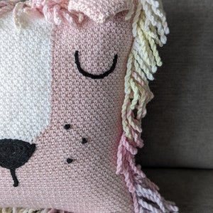 A crocheted pillow of a pink lion face on a grey couch