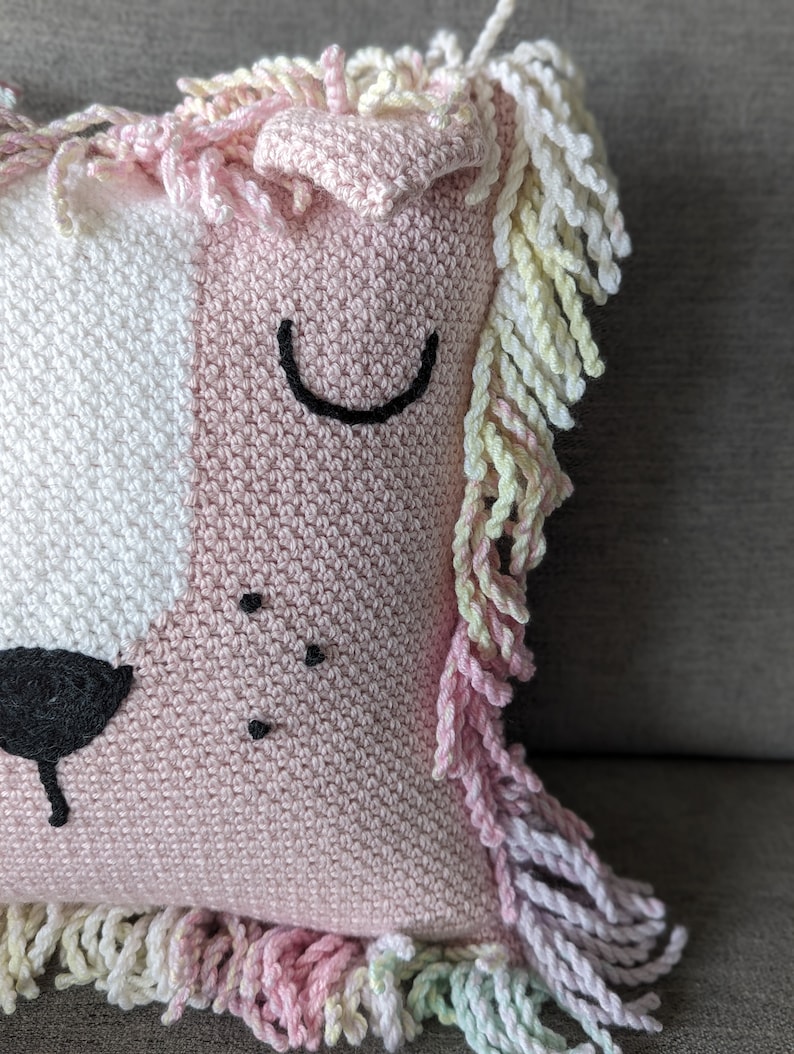 A crocheted pillow of a pink lion face on a grey couch