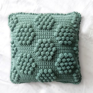 May include: A green crocheted cushion with a textured, raised pattern. The cushion is square shaped and has a fringed edge.