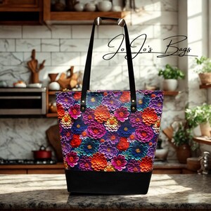 Op de afbeelding: Een tote bag met een levendig bloemenpatroon met paarse, oranje, rode en blauwe bloemen op het hoofdgedeelte en een effen zwarte onderkant. De tas heeft twee zwarte schouderbanden bevestigd met gouden klinknagels en toont de tekst Jo Jo's Bags.