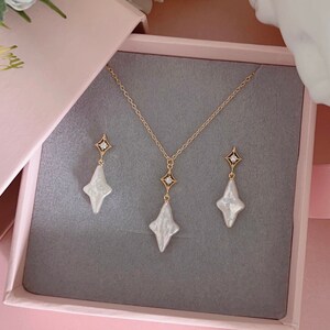 May include: A set of pearl jewelry in a pink box. The set includes a necklace and earrings. The necklace and earrings feature a gold chain with a white pearl pendant and a small diamond accent.