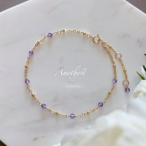 May include: Delicate gold bracelet featuring small, round amethyst beads. The bracelet has a delicate chain design with a clasp closure and adjustable length. The word "Amethyst" is written in cursive on the surface.