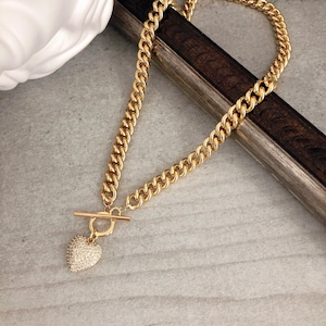 May include: Gold-toned chain necklace with a heart pendant. The necklace features a thick curb chain design and a toggle clasp. The heart pendant is encrusted with small, sparkling stones, creating a glamorous look. The necklace is displayed on a gray surface.