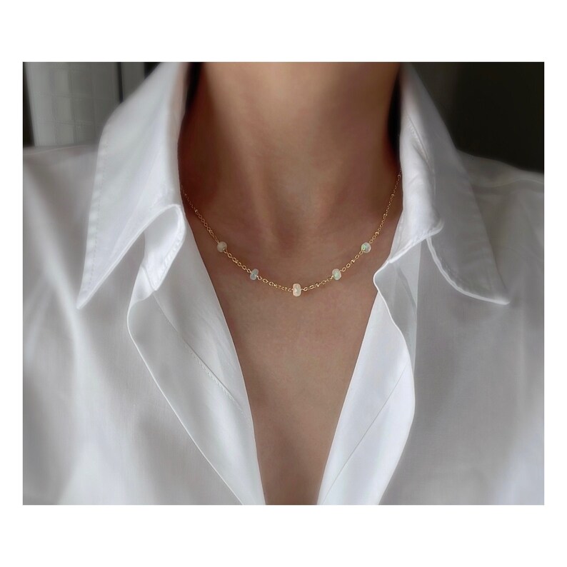 May include: A delicate gold chain necklace with small white opal stones. The necklace is worn by a person with a white shirt.