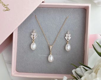 May include: A gold-toned jewelry set featuring a necklace and matching earrings, presented in a pink box. The necklace and earrings have a combination of clear crystals and pear-shaped pearls. The box is open, revealing the jewelry on a gray surface.