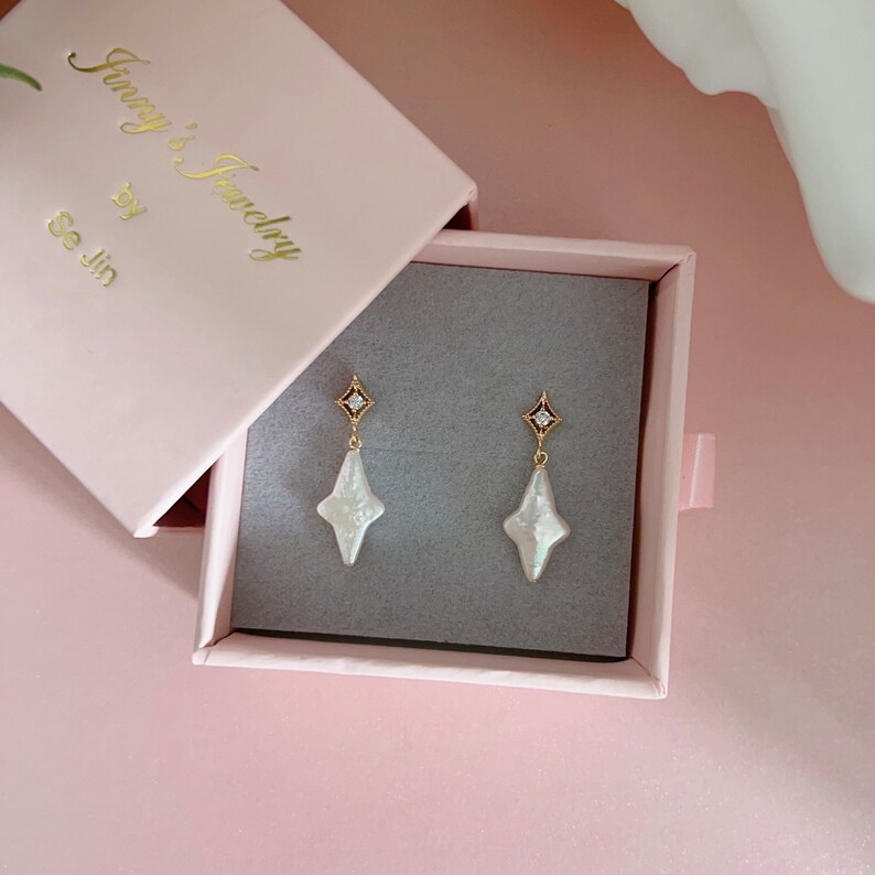 May include: A pair of gold earrings with a white pearl-like stone in the shape of a star. The earrings are in a small pink box with the text "Jenny's Jewelry by Se Jin" on the lid.