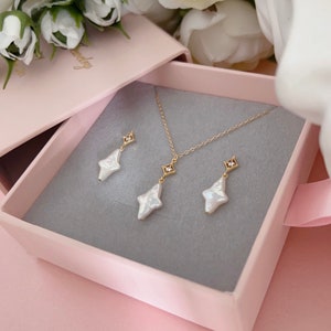 May include: A set of pearl and gold jewelry in a pink box. The set includes a necklace and earrings. The necklace has a delicate gold chain with a pearl pendant and a small diamond accent. The earrings are also pearl drop earrings with a diamond accent.