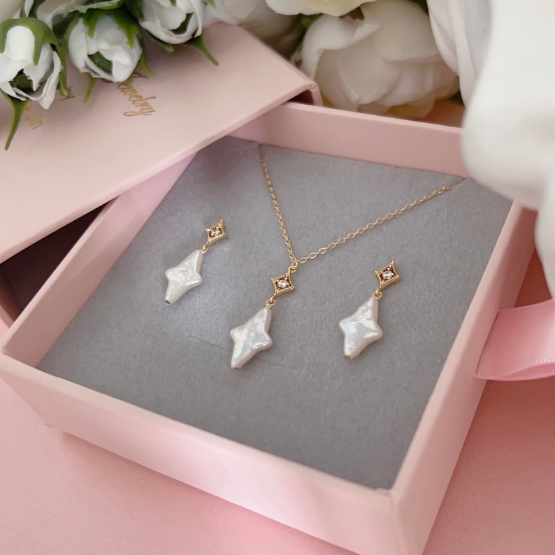 May include: A set of pearl and gold jewelry in a pink box. The set includes a necklace and earrings. The necklace has a delicate gold chain with a pearl pendant and a small diamond accent. The earrings are also pearl drop earrings with a diamond accent.