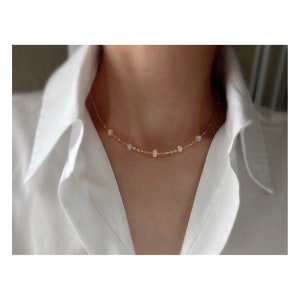 May include: A delicate gold chain necklace with small, iridescent opal beads. The necklace is worn by a person with a white shirt.