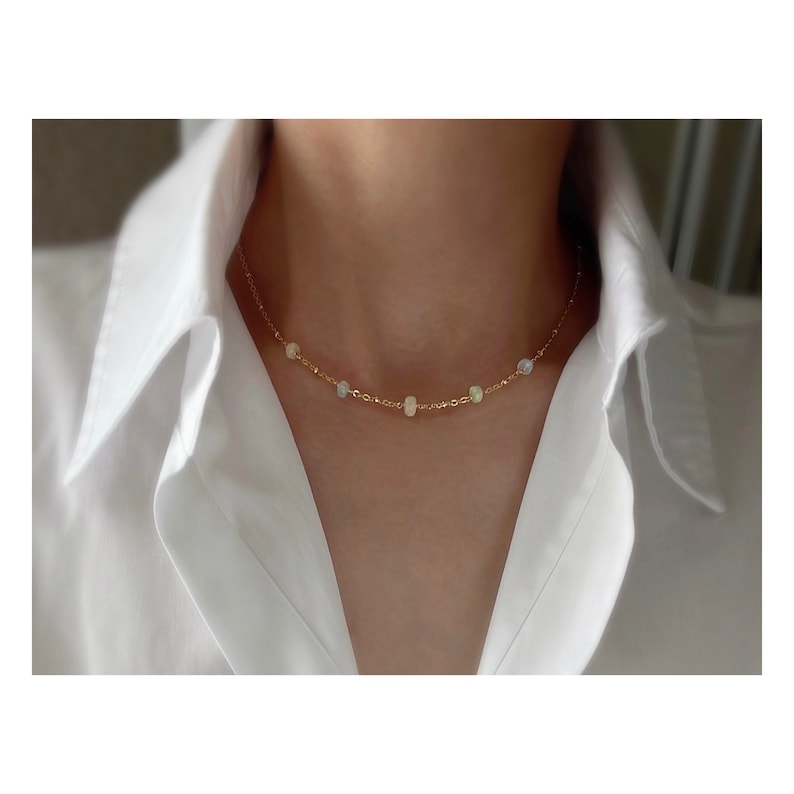May include: A delicate gold chain necklace with small, iridescent opal beads. The necklace is worn by a person with a white shirt.