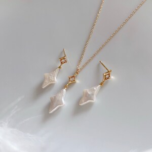 May include: A set of gold jewelry featuring a delicate necklace and matching earrings. The pendant and earrings are shaped like a diamond with a white pearl center and a small clear crystal at the top.