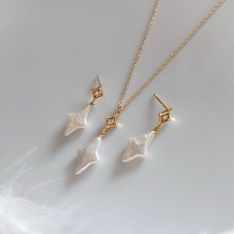 May include: A set of gold jewelry featuring a delicate necklace and matching earrings. The pendant and earrings are shaped like a diamond with a white pearl center and a small clear crystal at the top.