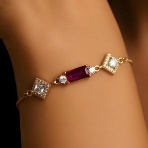 Pode incluir: Pulseira dourada com uma pedra retangular vermelha escura, ladeada por duas pedras redondas transparentes e duas cravações quadradas com diamantes. A pulseira é delicada e elegante, com uma corrente fina.