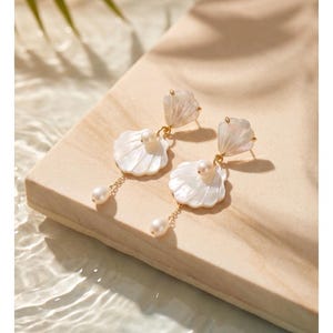 May include: A pair of drop earrings featuring white shell-shaped components with an iridescent finish. Each earring includes a small round pearl at the center of the shell and a delicate gold-tone chain dangling with a single teardrop pearl.