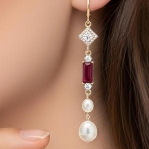 Peut inclure: Boucles d'oreilles pendantes dorées avec un dessus carré orné de diamants, un rubis rectangulaire et deux perles blanches. Les boucles d'oreilles sont suspendues à une chaîne délicate.