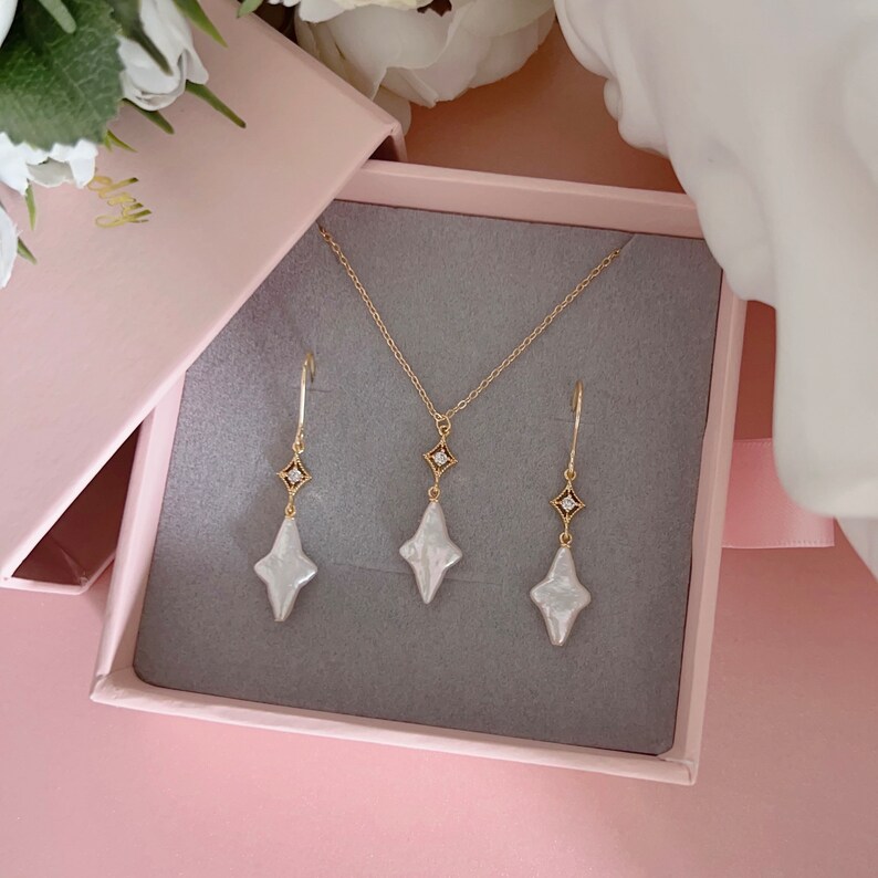 May include: A set of gold jewelry with white pearl accents. The set includes a necklace and a pair of earrings. The necklace and earrings feature a diamond-shaped pendant with a small diamond in the center.