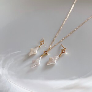 May include: A gold necklace and earrings set with a white pearl pendant and a small clear stone accent. The pendant and earrings are shaped like a diamond.