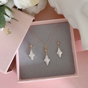 May include: A white pearl necklace and earrings set with a gold chain and a small diamond accent on each piece. The jewelry is displayed in a pink box.