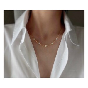 May include: A delicate gold chain necklace with small, round opal stones. The necklace is on a model wearing a white shirt.