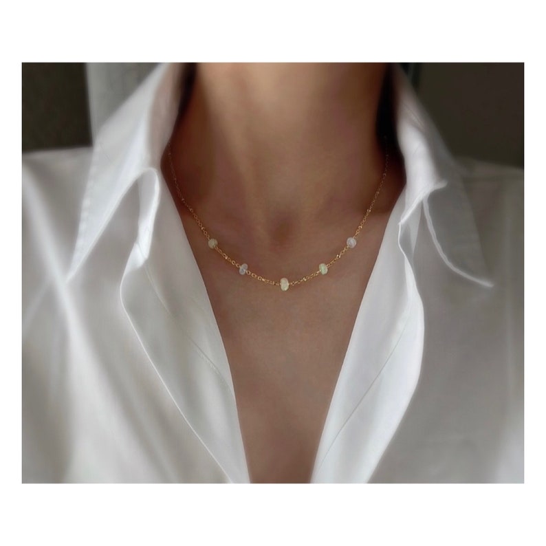 May include: A delicate gold chain necklace with small, round opal stones. The necklace is on a model wearing a white shirt.