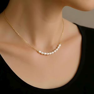 May include: A gold necklace with a string of small, white pearls. The pearls are arranged in a gentle curve. The necklace is worn around the neck, with the gold chain visible. A simple and elegant piece of jewellery.