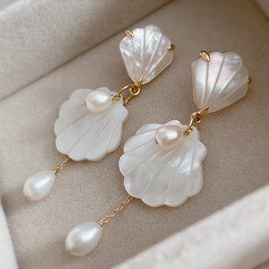 Peut inclure: Une paire de boucles d'oreilles pendantes composées d'éléments en forme de coquillage blanc irisé et de perles blanches lustrées. Chaque boucle présente une pièce supérieure en coquillage reliée par un anneau doré à une pièce plus grande.